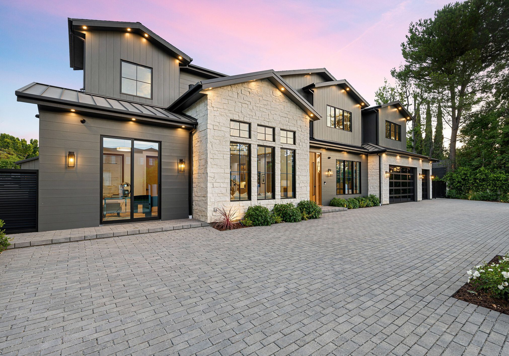 Modern luxury home exterior with large windows, stone facade, and spacious driveway at sunset.