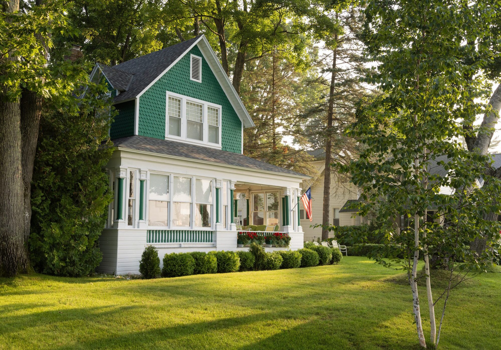 With an American flag proudly displayed, a corner view of a Lake Michigan seasonal summer cottage that is painted green and white. Built in the late 1800's, the house is surrounded by a lush lawn, trees and bushes. It is the perfect escape for vacation, rest and relaxation.